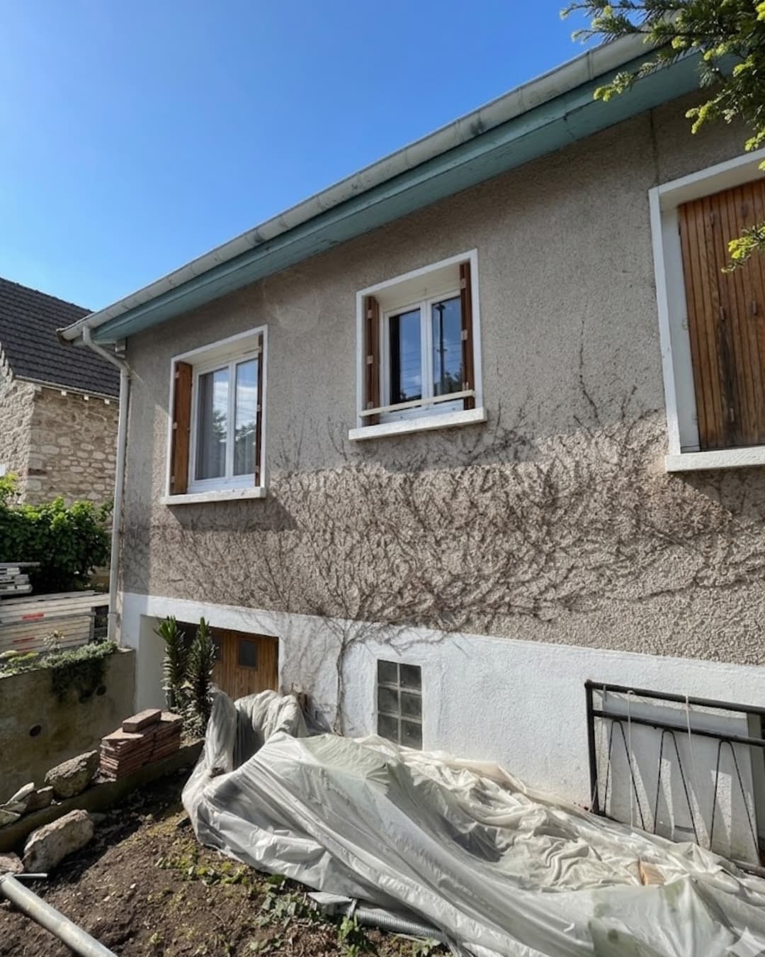 Vue d'ensemble de la façade avant d'une maison à Taverny, montrant un enduit rugueux d'origine avec des traces de végétation grimpante sèche et un débord de toit bleu clair.